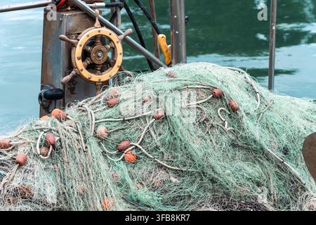 Ein großes Fischernetz ruht auf einem Bootsdeck neben einem Steuerrad und ruhigem Wasser. Stockfoto