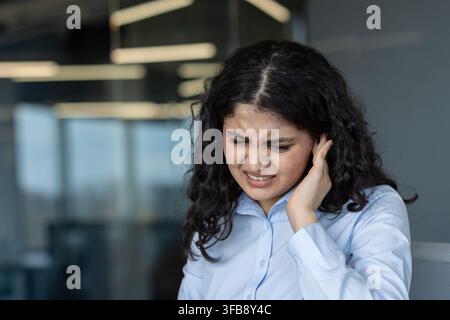 Junge Geschäftsfrau, die im Büroumfeld starke Ohrschmerzen und Unbehagen erlebt, ihr schmerzendes Ohr mit einer Grimasse haltend, was auf Gesundheitsprobleme oder Stress hinweist, die ihre Konzentration beeinträchtigen Stockfoto