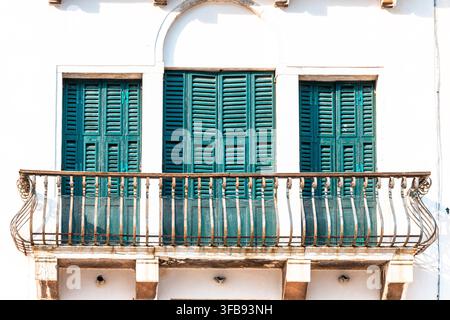 Eine sonnenbeschienene weiße Gebäudefassade zeigt türkisfarbene Fensterläden und einen schmiedeeisernen Balkon. Stockfoto