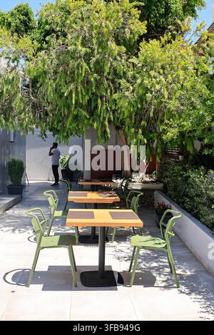 Oia, Santorin, Griechenland - 4. August 2026: Leere Café-Tische und grüne Stühle unter einem schattigen Baum Stockfoto