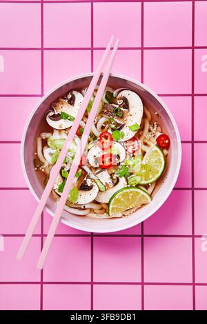 Asiatische Nudelsuppe mit frischem Gemüse, serviert in einer rosa Schüssel mit rosa Essstäbchen. Der Hintergrund zeigt rosa Fliesen und erzeugt eine weiche, stilvolle und Stockfoto