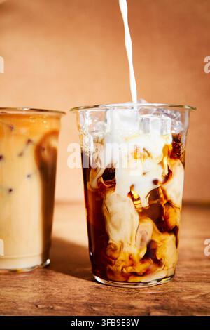 Das Mischen von Kaffee und Milch in einem Glas. Milch wird von oben in ein Kaffeeglas mit Eiswürfeln gegossen. Warme, gemütliche Beleuchtung. Stockfoto