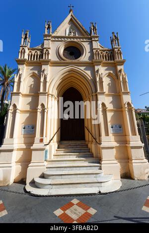 Neogotische rumänisch-orthodoxe Kirchenfassade in Sanremo, Italien, mit einem Rosettenfenster, spitzbögenförmigem Eingang, cremefarbenem Stein und einem hohen Kreuz unter blauem Himmel. Stockfoto