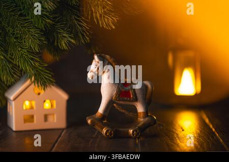 Miniaturspielzeug mit Schaukelpferd, das eine festliche weihnachtsszene auf einem Holztisch dekoriert, mit Keramikhaus, grünen Kiefernzweigen und warmer Atmosphäre Stockfoto