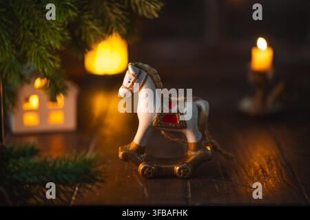 Miniaturspielzeug mit Schaukelpferd, das eine festliche weihnachtsszene auf einem Holztisch dekoriert, mit Keramikhaus, grünen Kiefernzweigen und warmer Atmosphäre Stockfoto