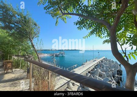 Hafen auf der Schwarzmeerinsel. Kleiner Hafen mit Booten, Betonmolle und ruhiger blauer See, der durch grüne Bäume an einem sonnigen Sommertag zu sehen ist. Küste Stockfoto