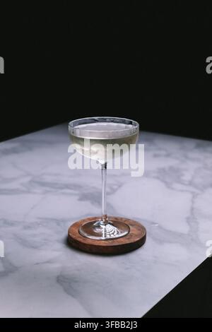 Blasser Cocktail in einem genagelten Coupé-Glas auf einem Holzuntersetzer auf weißem Marmor vor dunklem Hintergrund, stimmungsvoller Getränkefotografie Stockfoto