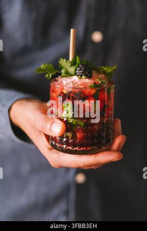 Hand hält einen Blackberry-Cocktail mit zerstoßenem Eis, Minze und einem Papierstrohhalm in einem Rocks-Glas, Nahaufnahme Stockfoto