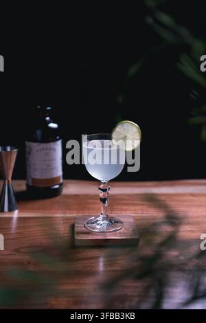 Blasser violetter Cocktail mit einer Limetten-Scheibe in einem Vintage-Weinglas auf einem Holzuntersetzer in einer dunklen, stimmungsvollen Hausbar Stockfoto