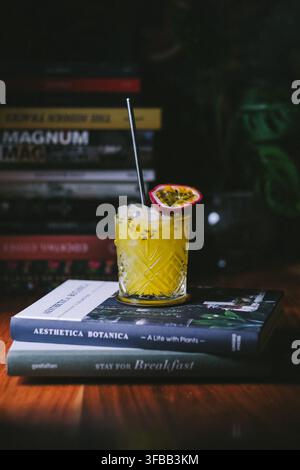 Gelber Passionsfrucht-Cocktail in einem geschliffenen Kristallglas auf einem Stapel Bücher vor einem Bücherregal, stimmungvolle Getränkefotografie Stockfoto