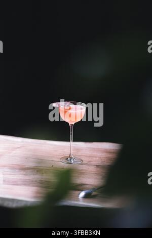 Pinkfarbener Cocktail mit einer essbaren Blumengarnitur in einem Coupé-Glas auf einem hellen Holzbrett vor einem dunklen Hintergrund Stockfoto