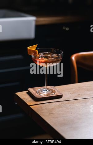Whisky-Cocktail mit klarem Eiswürfel und Orangenschale in einem Coupé-Glas auf einem Holzuntersetzer in einer dunklen Küche, stimmungsvolle Craft-Cocktail-Fotografie Stockfoto