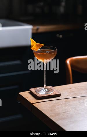 Bernstein-Craft-Cocktail in einem Coupé-Glas mit Orangenschalen-Twist auf einem Holzuntersetzer in einer dunklen modernen Küche, stimmungsvolle Hausbarkeeper-Getränkefotografie Stockfoto