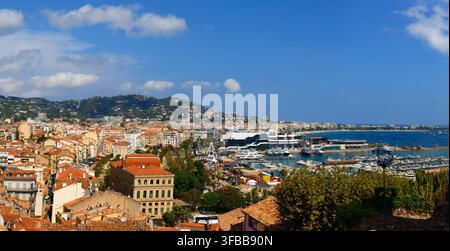 Blick auf die Marina entlang der Küste von Cannes, Frankreich, mit Segelbooten und Yachten, die neben einer historischen Hügelstadt mit einem alten Steinturm in der Ferne vertäut sind Stockfoto
