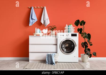 Moderne Waschraum-Innenarchitektur, Konzept für Heimorganisation und Reinigung, Waschmaschine mit Waschmitteln und Pflanzen auf orangefarbenem Wandhintergrund Stockfoto