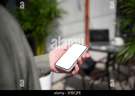 Nahaufnahme eines reifen Freelancers, der ein Smartphone mit leerem weißem Bildschirm in der Nähe eines Laptop-Arbeitsplatzes hält, mobile App-Vorlage, Remote-Arbeit und digitale Technologie Stockfoto