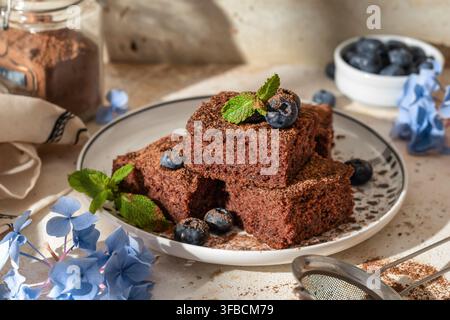 Schokoladenbrowniequadrate, die mit Minzblättern verziert sind, im hellen Morgenlicht mit harten Schatten auf einem rustikalen Küchentisch. Horizontal Stockfoto