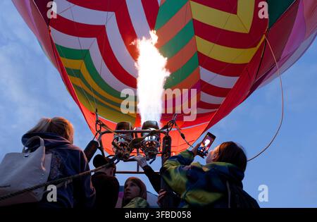 Niedrigwinklige Aufnahme von Menschen, die zuschauen und Fotos machen, während eine starke Propaneflamme einen farbenfrohen Heißluftballon entzündet. Stockfoto