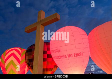 Niedrigwinklige Perspektive eines großen steinernen Kreuzdenkmals auf dem Piłsudski-Platz, das sich vor leuchtenden Heißluftballonen und einem dunkelblauen Abendhimmel abhebt. Stockfoto