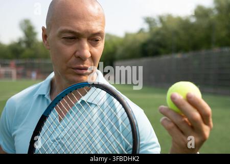 Konzentrierter reifer kaukasischer Tennisspieler, der einen Tennisball betrachtet, während er einen Schläger auf einem Außenplatz vor dem Training oder Match hält. Stockfoto