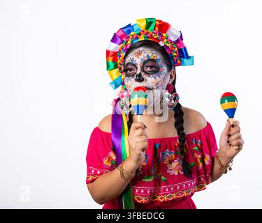 Fröhliche Frau mit traditionellem Tag-der-Toten-Make-up und farbenfroher mexikanischer Kleidung, die lebhafte Maracas vor weißem Hintergrund hält. Stockfoto