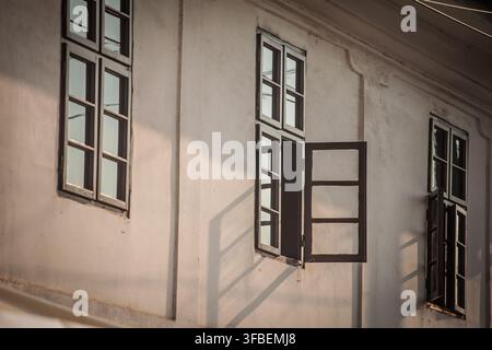 Offene holzgerahmte Fenster an einem alten Gebäude werfen lange geometrische Schatten auf eine abgenutzte Putzwand und betonen traditionelle Architektur, Alter und n Stockfoto