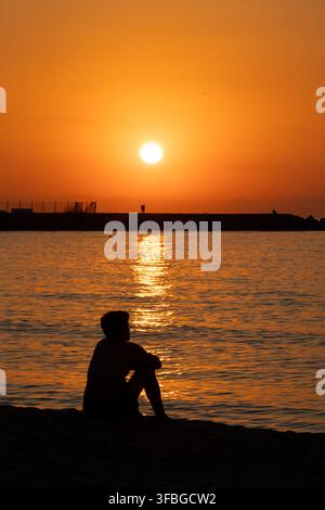 Ein einsamer Mann sitzt als Silhouette am Ufer und beobachtet einen lebhaften orangefarbenen Sonnenuntergang über dem Meer, während die tiefstehende Sonne eine lange goldene Spiegelung erzeugt Stockfoto