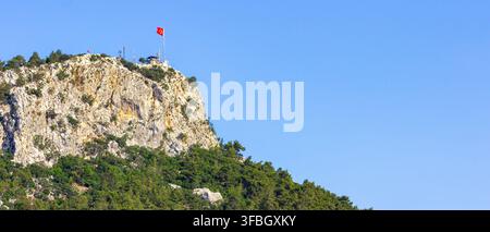 Panoramische kinematografische Ansicht des Berges Calis (Idyros) in Kemer, Türkei, mit der türkischen Flagge auf dem Gipfel und weitem blauem Himmel über felsigen Klippen und Wald Stockfoto
