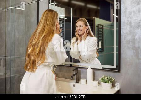 Lächelnde junge Frau steht im Badezimmer, schaut in den Spiegel und genießt ihre morgendliche Hautpflegeroutine Stockfoto