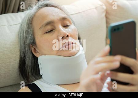 Nahaufnahme einer reifen Patientin mit einer Halsverletzung, die einen medizinischen Halskragen trägt und auf ein Mobiltelefon schaut, um einen Arzt online zu konsultieren oder zu überprüfen Stockfoto