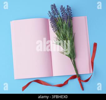 Öffnen Sie das Notizbuch mit leeren rosafarbenen Seiten, Blumenstrauß aus Lavendelchen auf blauem Hintergrund, Draufsicht, flacher Laie Stockfoto