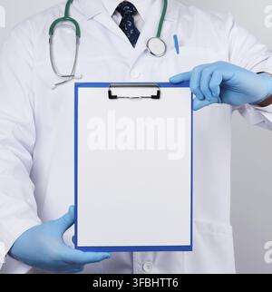 Hausarzt in einem weißen Mantel, blaue Latexhandschuhe, ein Stethoskop, das an seinem Hals hängt und einen Papierhalter mit einem weißen leeren Blatt hält, Platz zum Schreiben eines Stockfoto