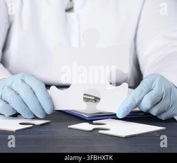 Der Arzt sitzt am Tisch und hält ein weißes Papierpuzzle. Lösungsbegriff, fehlendes Stück Stockfoto