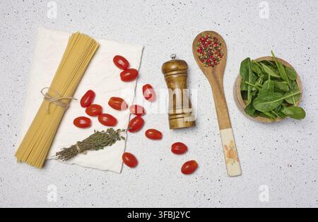 Hölzerne Schüssel mit Spinat, Kirschtomaten auf einem Holzteller, Löffel mit Gewürzen, ungekochte Spaghetti mit Garnbindung Stockfoto