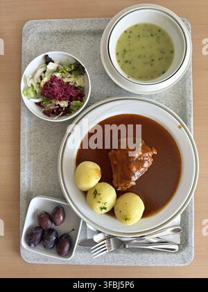 Auf einem Teller liegt ein Stück Fleisch in dicker brauner Sauce sowie drei gekochte Kartoffeln, garniert mit Petersilie. Zusätzlich eine Schüssel cremige grüne Kräutersuppe a Stockfoto