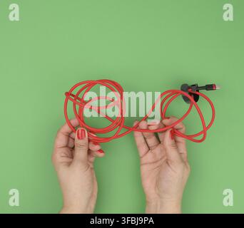 Weibliche Hand hält verdrilltes Kabel zum Laden von mobilen Geräten in einer roten Textilverpackung auf grünem Hintergrund, Nahaufnahme Stockfoto
