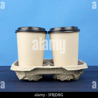 Zwei braune Einwegbecher aus Papier mit einem Plastikdeckel stehen im Tablett auf einem Holztisch, blauer Hintergrund, Behälter zum Mitnehmen Stockfoto