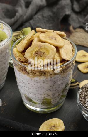Zwei Gläser Chiasamenpudding, eines mit Kiwi-Würfel und das andere mit Müsli und Bananenchips, befinden sich auf einer dunklen Oberfläche mit Zutaten Stockfoto