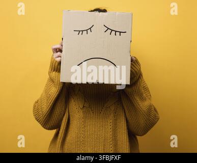 Eine Frau in einem braunen Pullover mit einer Kiste auf dem Kopf steht auf einem gelben Hintergrund. Trauriges Grimassen, Stress- und Depressionskonzept. Psychische Gesundheit Stockfoto