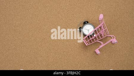 Rosa Einkaufswagen und schwarzer runder Wecker innen auf braunem Hintergrund. Zeit ist Geld, Banner Stockfoto