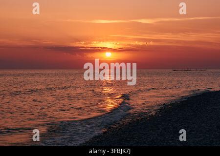Ein atemberaubender Sonnenuntergang wirft ein warmes orangefarbenes Licht über das ruhige Meer und die Kiesküste, mit sanften Wellen und fernen Hügeln, aufgenommen bei Dionisiou B Stockfoto