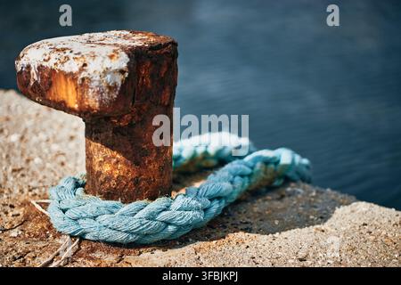 Nahaufnahme eines stark verwitterten und rostigen Metall-Landezapfens, der mit einem hellblauen geflochtenen Seil an einem Betonpier befestigt ist, vor einem ruhigen dunklen Hintergrund Stockfoto
