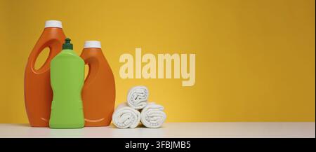 Gedrehte Lappen zum Wischen von Böden und Kunststoffflaschen für chemische Flüssigprodukte, Gel, Reinigungsmittel auf einem weißen Tisch, gelber Hintergrund. Speicherplatz kopieren Stockfoto