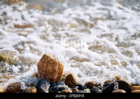 Nahaufnahme eines nassen braunen Kieselsteins, der zwischen Kieselsteinen an einer Meeresküste ruht, während weißer Schaum von anrollenden Ozeanwellen über den Strand spült Stockfoto