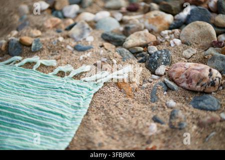 Nahaufnahme eines gestreiften grün-weißen Strandtuchs mit Fransen, das auf sandigem Boden liegt, umgeben von kleinen Steinen und glatten Meereskieseln. Stockfoto