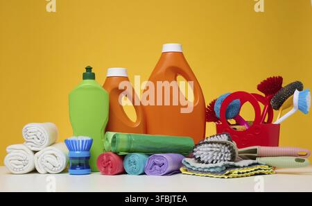 Orangefarbene Kunststoffflaschen mit flüssigen Produkten und Müllsäcken, Bürsten und Textillappen zum Waschen von Böden auf einem weißen Tisch, gelber Hintergrund Stockfoto
