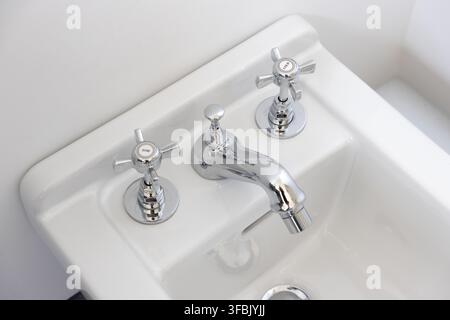 Vintage-Chrom-Wasserhahn mit separaten Warm- und Kaltwasserhähnen auf einem weißen Keramikwaschbecken Stockfoto