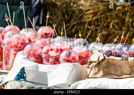 Portionierte frische Wassermelonenstücke und Beeren, verpackt in Plastik-To-Go-Bechern mit Holzspießen, die auf einem Marktstand im Freien angeordnet sind. Stockfoto