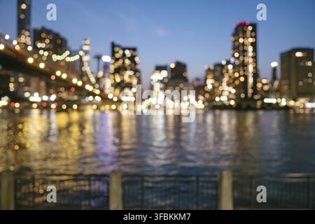 Unscharfe Stadtlichter und Brückenlampen leuchten über dem East River in dieser träumerischen New Yorker Dämmerungsszene. Die Reflexionen erzeugen ein abstraktes, farbenfrohes Stockfoto