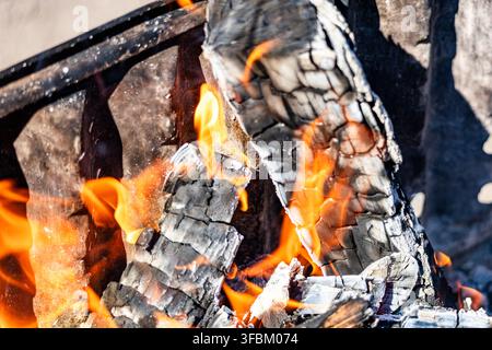 Horizontale Makroaufnahme von leuchtend orangefarbenen Feuerflammen, die schwarze verkohlte Holzscheite in einem Grillrahmen im Freien bei hellem Tageslicht verbrennen. Stockfoto
